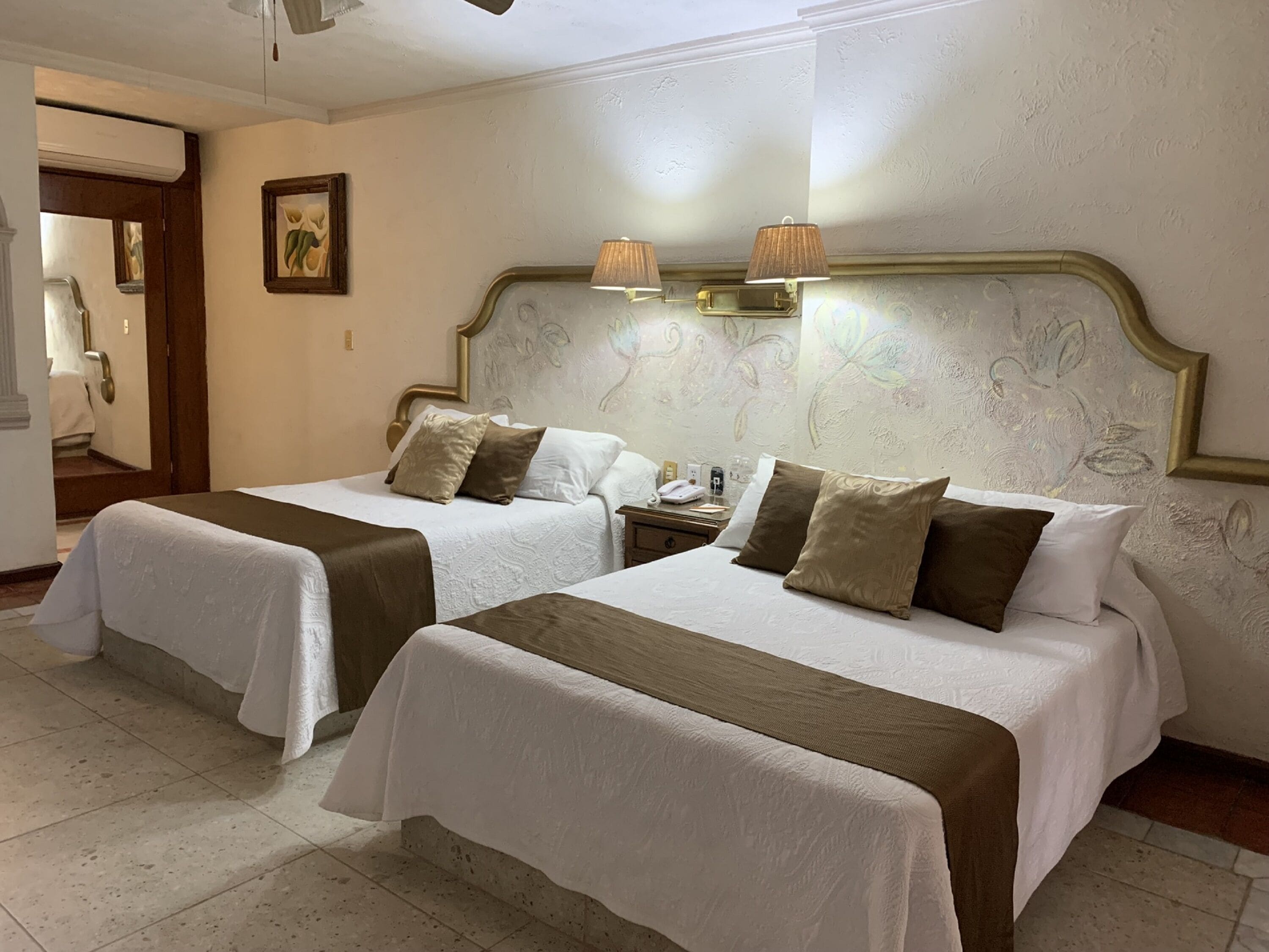 A hotel room with two beds, each with white linens and brown headboards. There are wall-mounted lamps above the beds and a painting on the wall.
