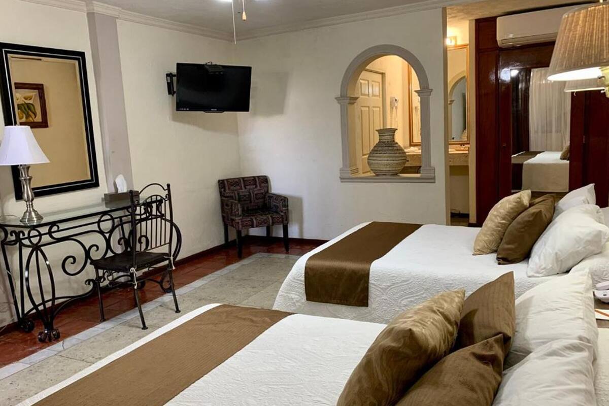Habitación de hotel con dos camas, televisor, escritorio con silla y un espejo decorativo.