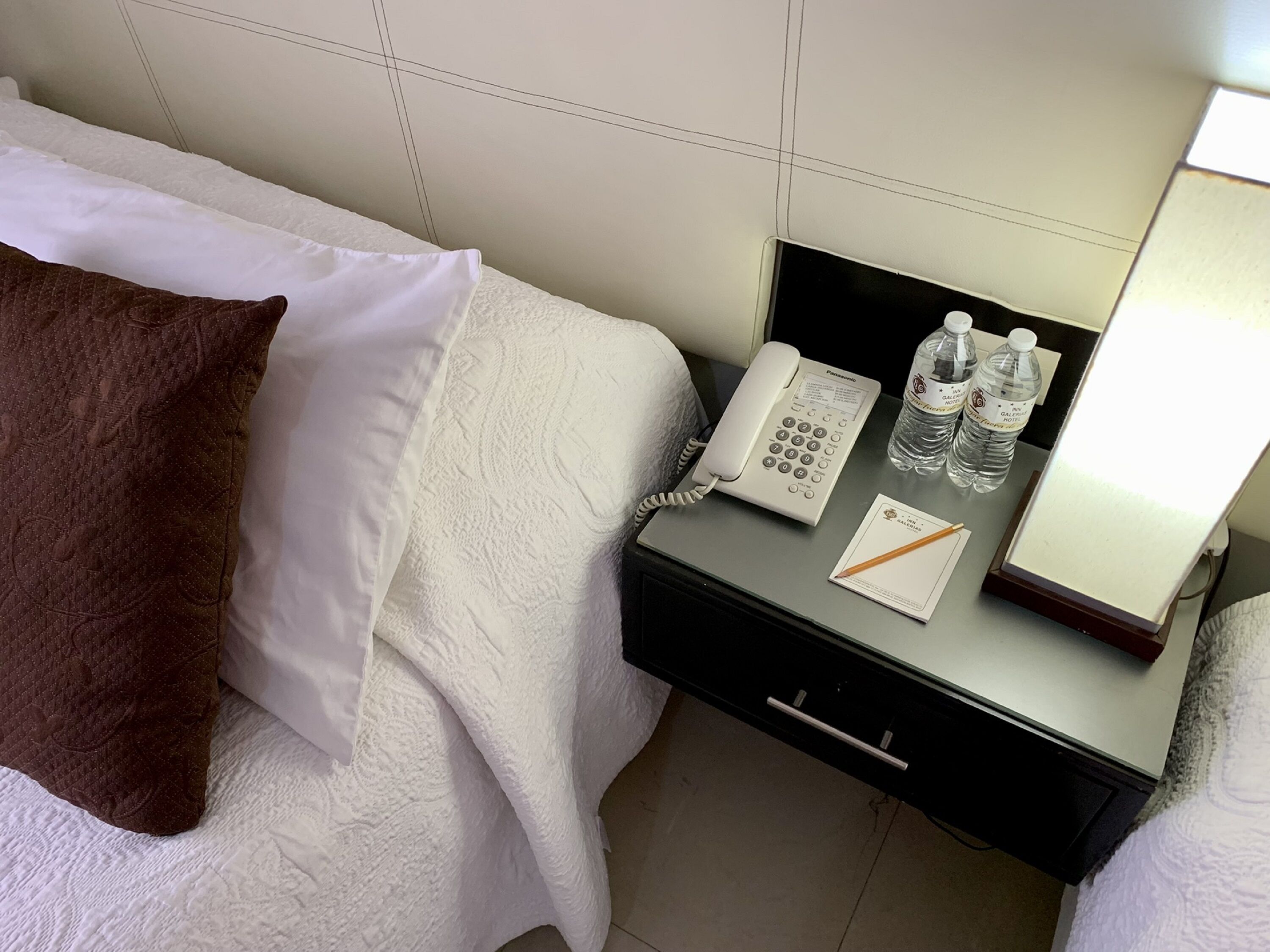 A hotel room with a bed, a nightstand with a phone, water bottles, and a notepad.