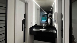 A modern hotel corridor with a black countertop sink, a trash bin, and a mirror reflecting a person. - Uniclass Hotel Lapa (São Paulo)