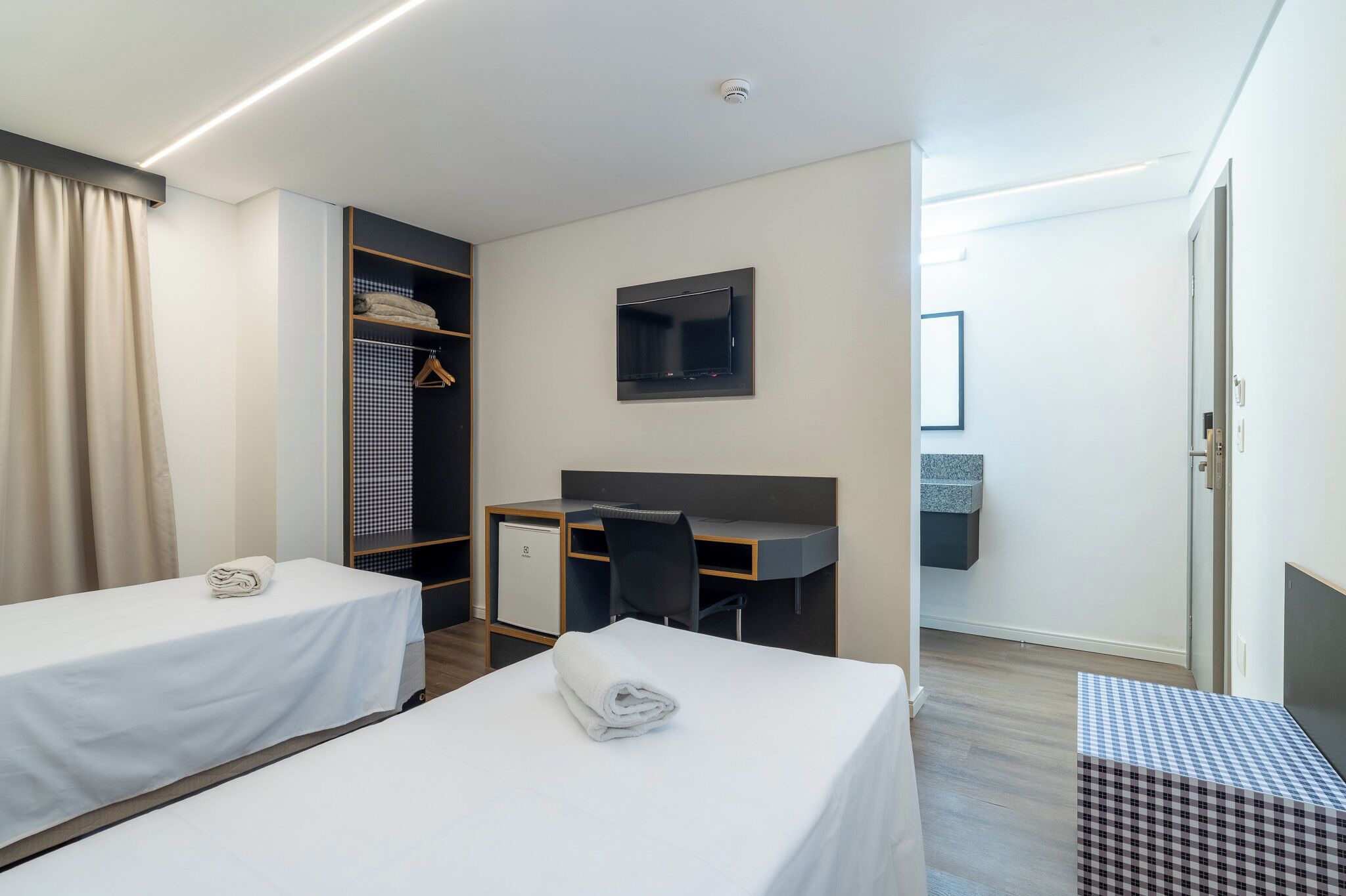 A hotel room with two beds, a desk, a wardrobe, a TV, and a small kitchenette.