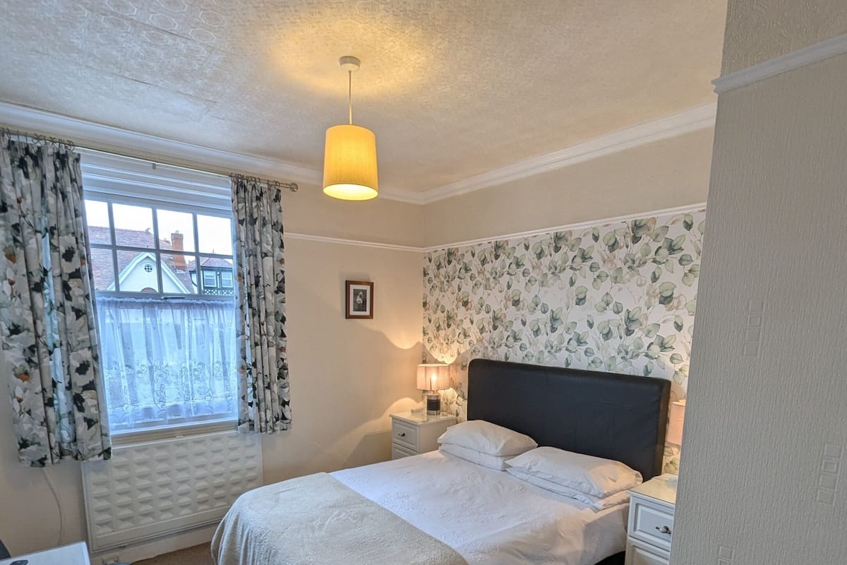 A bedroom with a bed, bedside tables, a lamp, a radiator, a window with curtains, and a view of buildings outside.