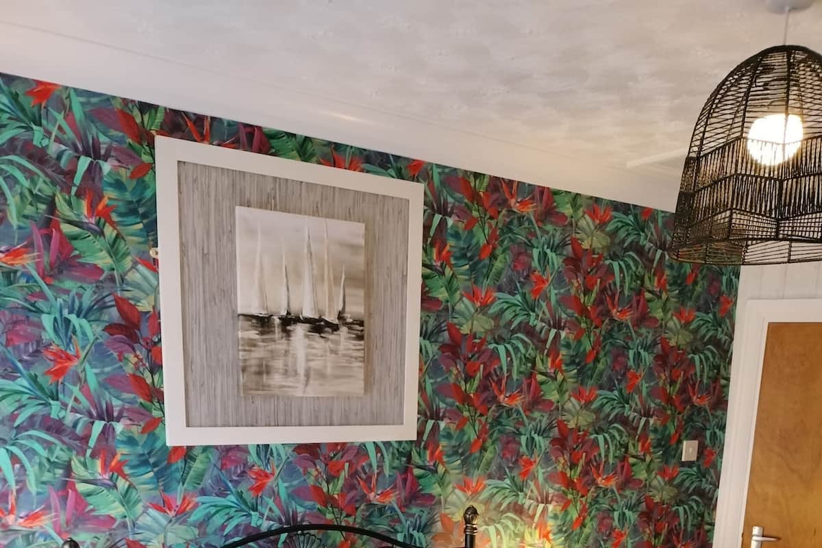 A neatly made bed with white and pink bedding, a framed picture above it, and a tropical wallpaper.
