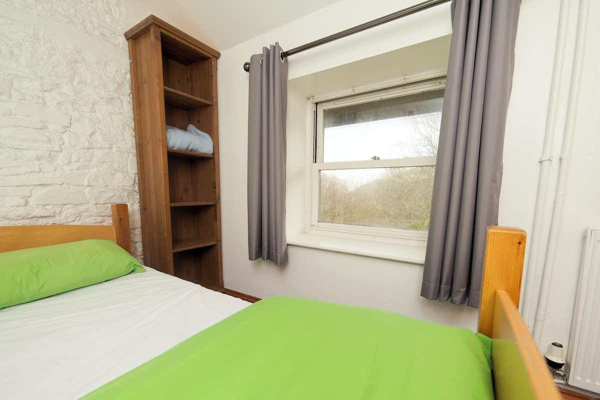 A bedroom with a wooden wardrobe, a window with grey curtains, and a bed with green bedding.
