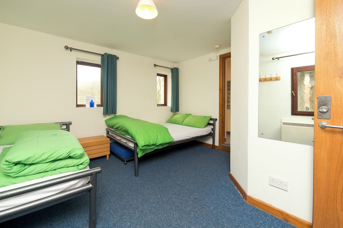 A room with two beds, a wooden door, a window with curtains, and a light fixture.