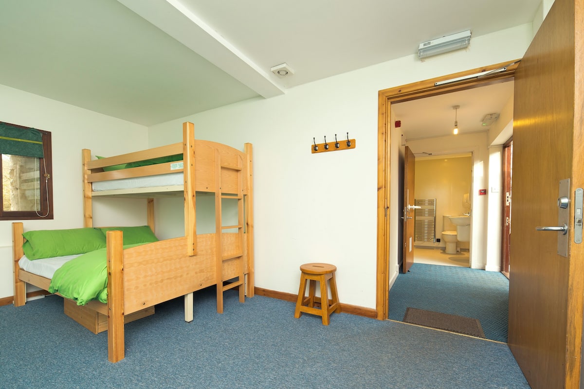 A room with bunk beds, a small stool, a door, and a visible bathroom in the background.