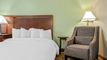 A hotel room with a large bed, a wooden headboard, a grey armchair, a small table, and a lamp.