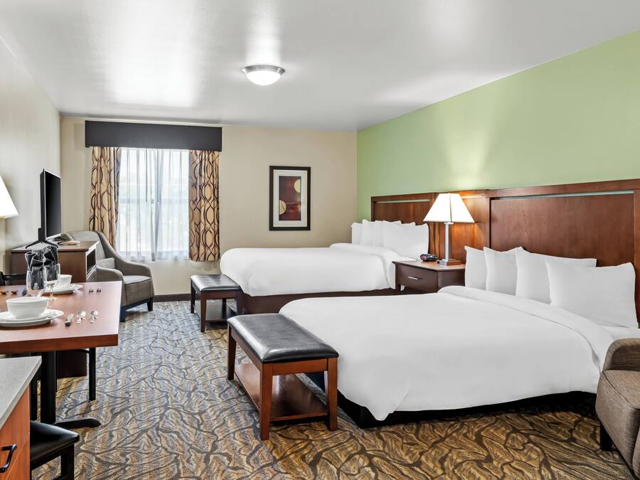 A hotel room with two beds, a desk with a chair, a small table, a lamp, and a window with curtains.