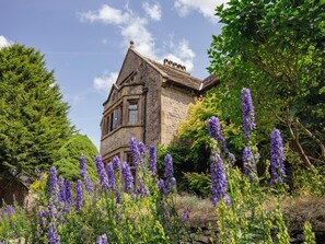 A historic stone building with a prominent tower, surrounded by lush greenery and purple flowers. - YHA Hartington Hall - Hostel (Buxton)