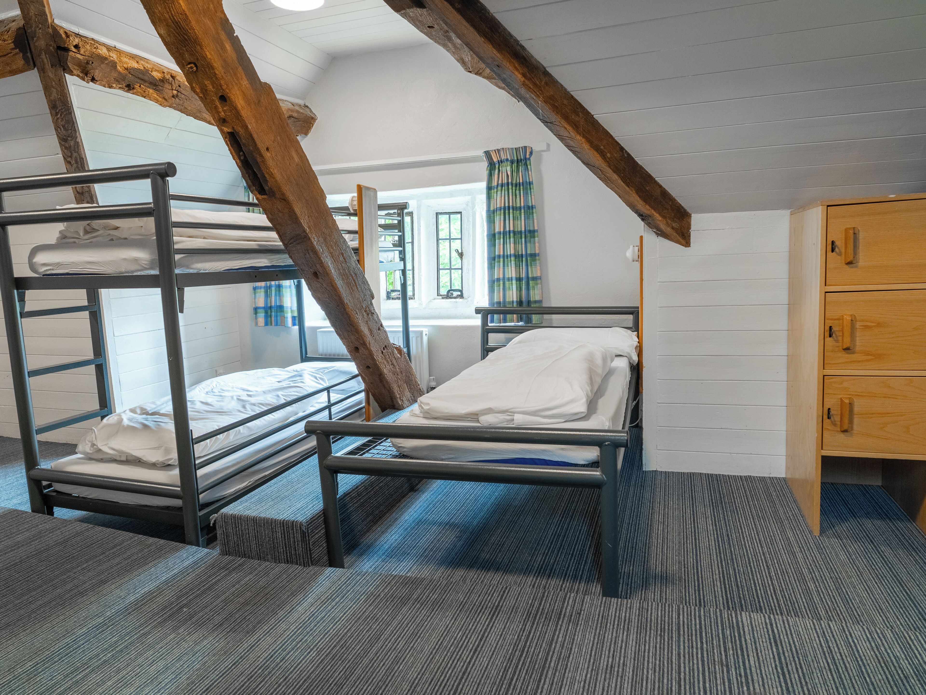 A dormitory room with bunk beds, a wooden wardrobe, and a window with curtains.