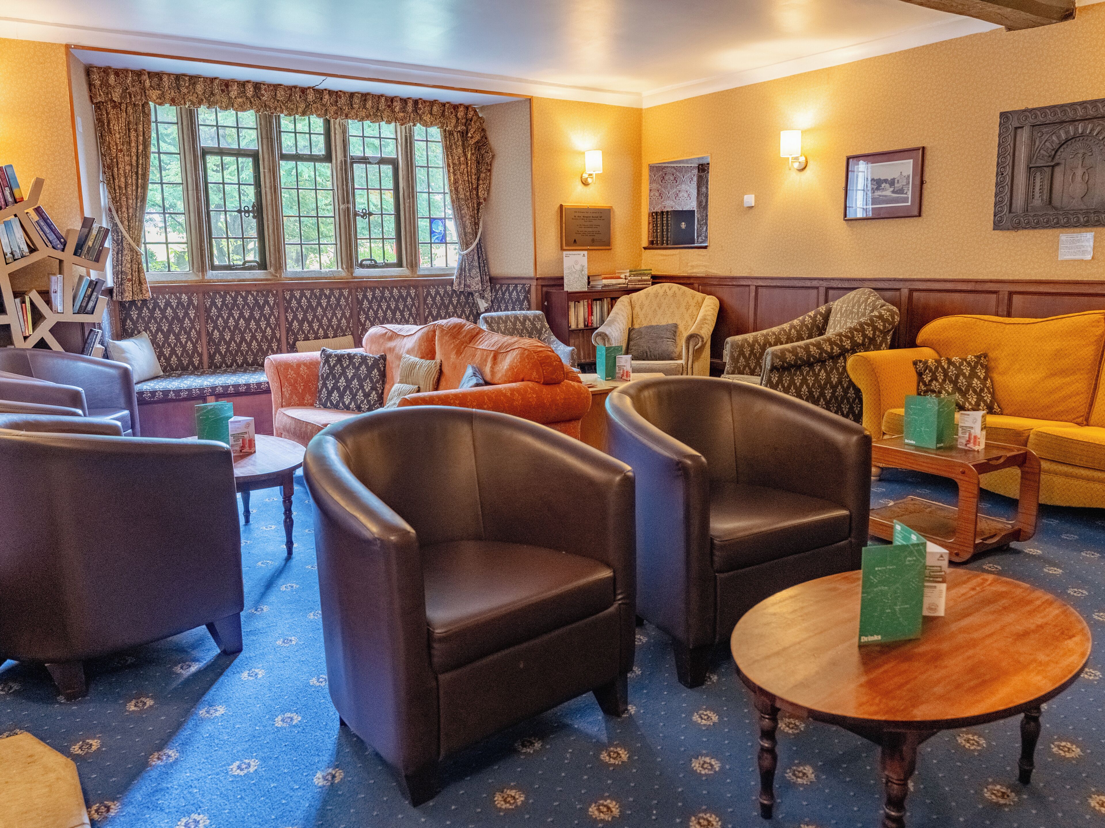 A cozy hotel room with a blue carpet, leather armchairs, a yellow sofa, and a bookshelf.