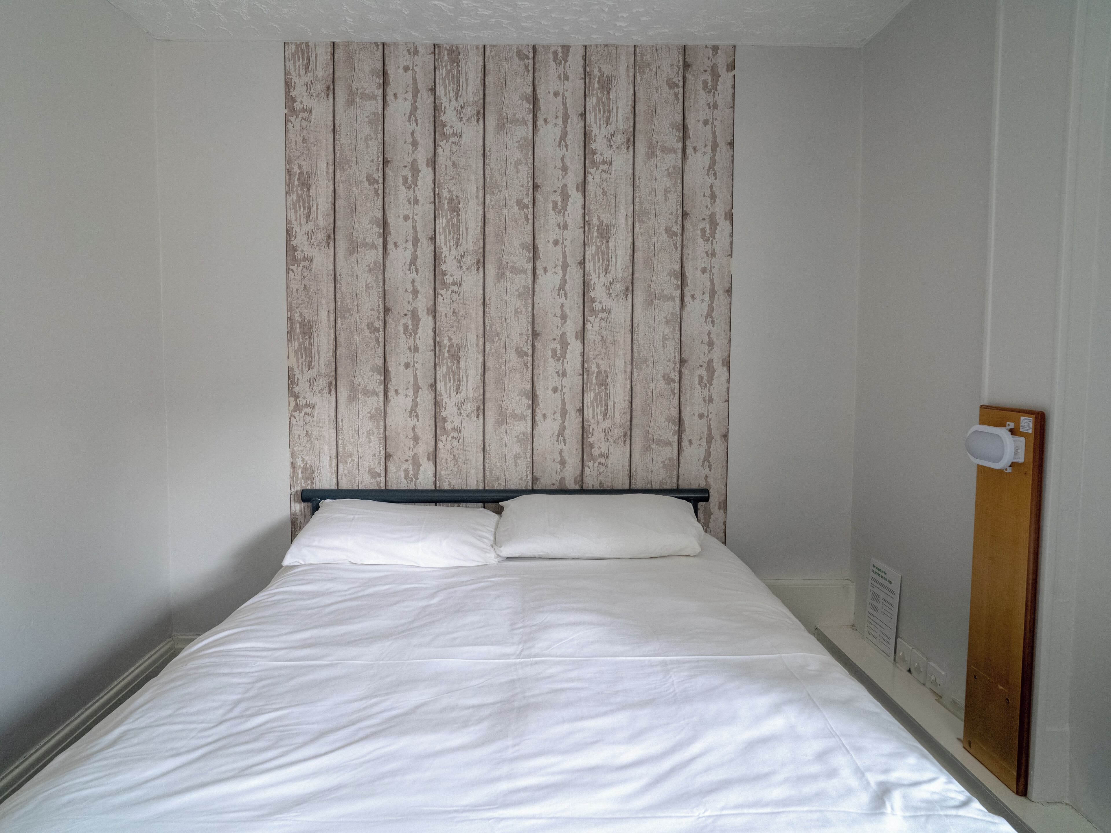 Una cama individual con ropa de cama blanca en una habitación con cabecera de paneles de madera y puerta de madera.