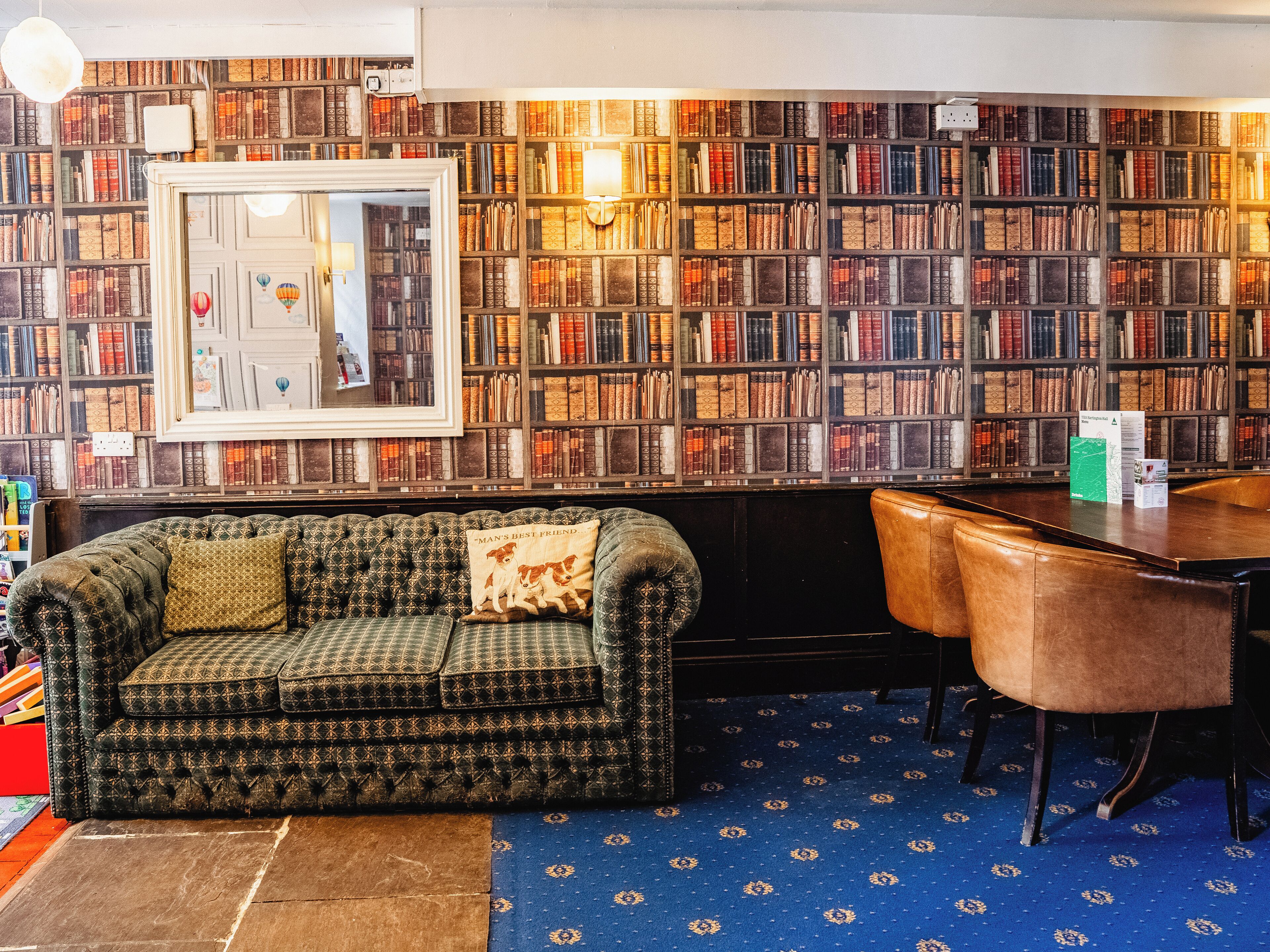 Ein gemütliches Arbeitszimmer mit einem grün gemusterten Sofa, einem Holztisch und einem Bücherregal voller Bücher.