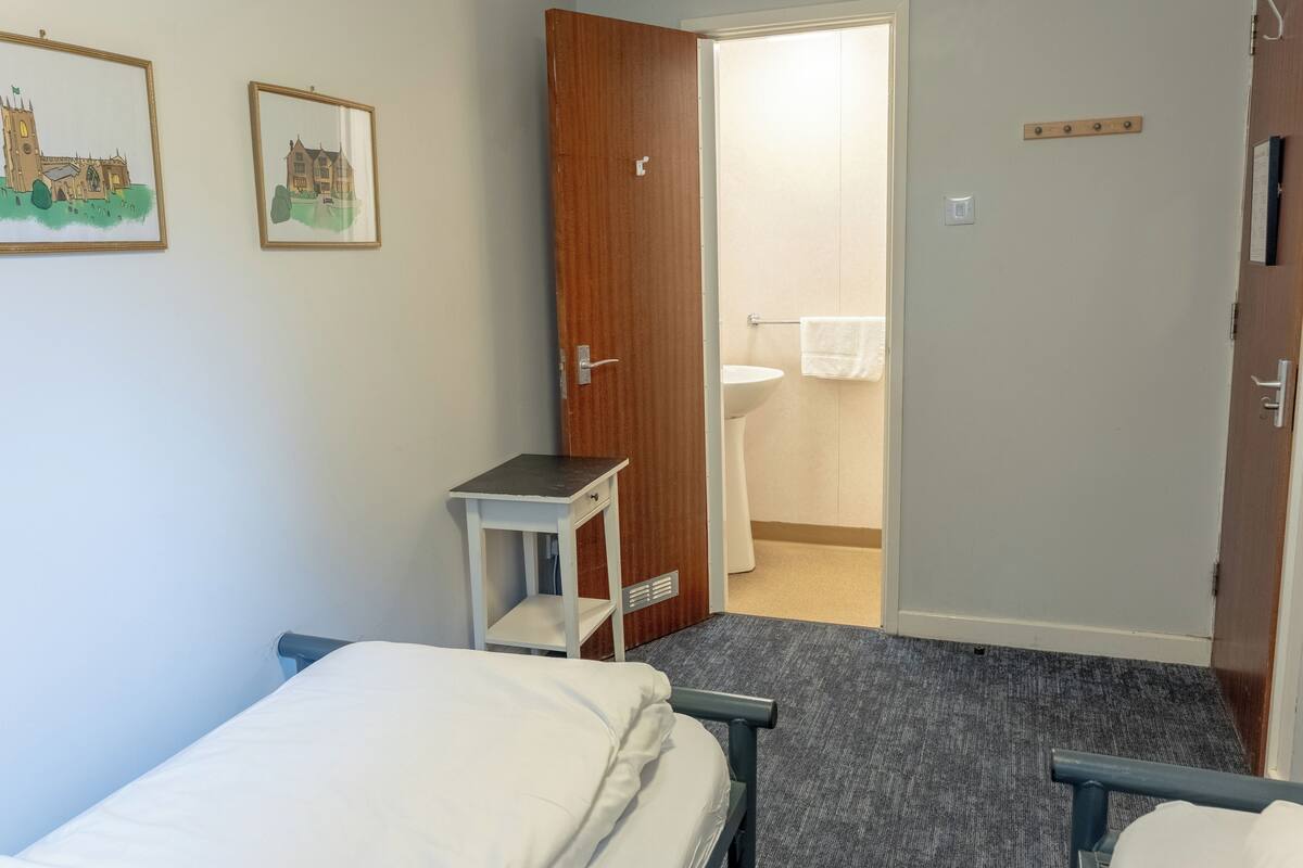 A small, clean room with a bed, a bedside table, a wooden door, and a bathroom visible through an open door.
