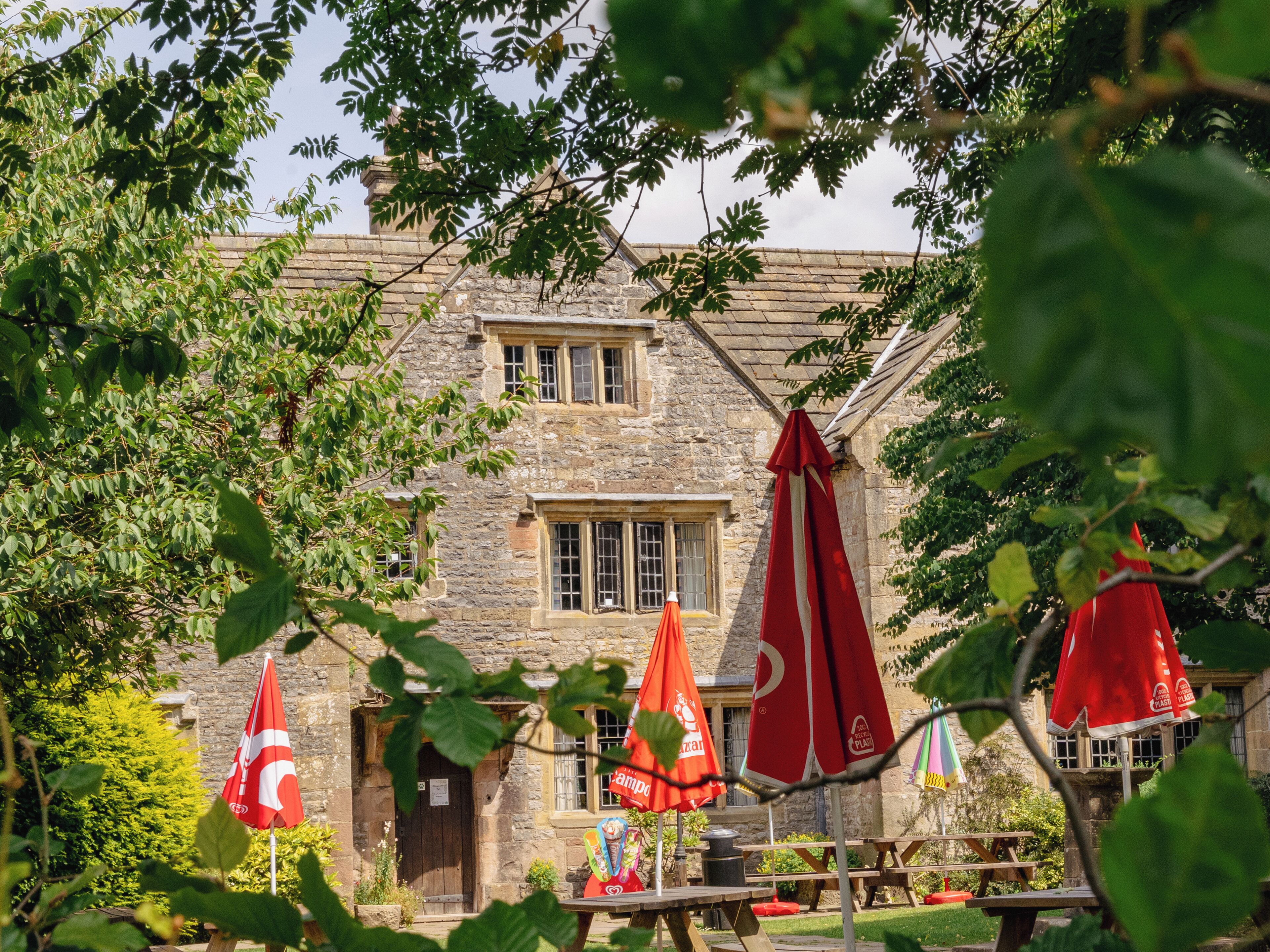 A stone building with multiple windows and a door, surrounded by greenery and red umbrellas.
