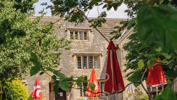 A stone building with multiple windows and a door, surrounded by greenery and red umbrellas.