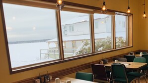 A dining area with wooden tables and chairs, large windows with a view of a snowy landscape, and pendant lighting. - Pickle Lake Hotel (Pickle Lake)