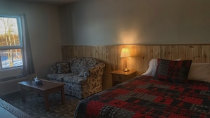A cozy room with a wooden headboard, a bed with a red and black quilt, a patterned armchair, a small table with a lamp, and a window with a view of trees. - Pickle Lake Hotel (Pickle Lake)
