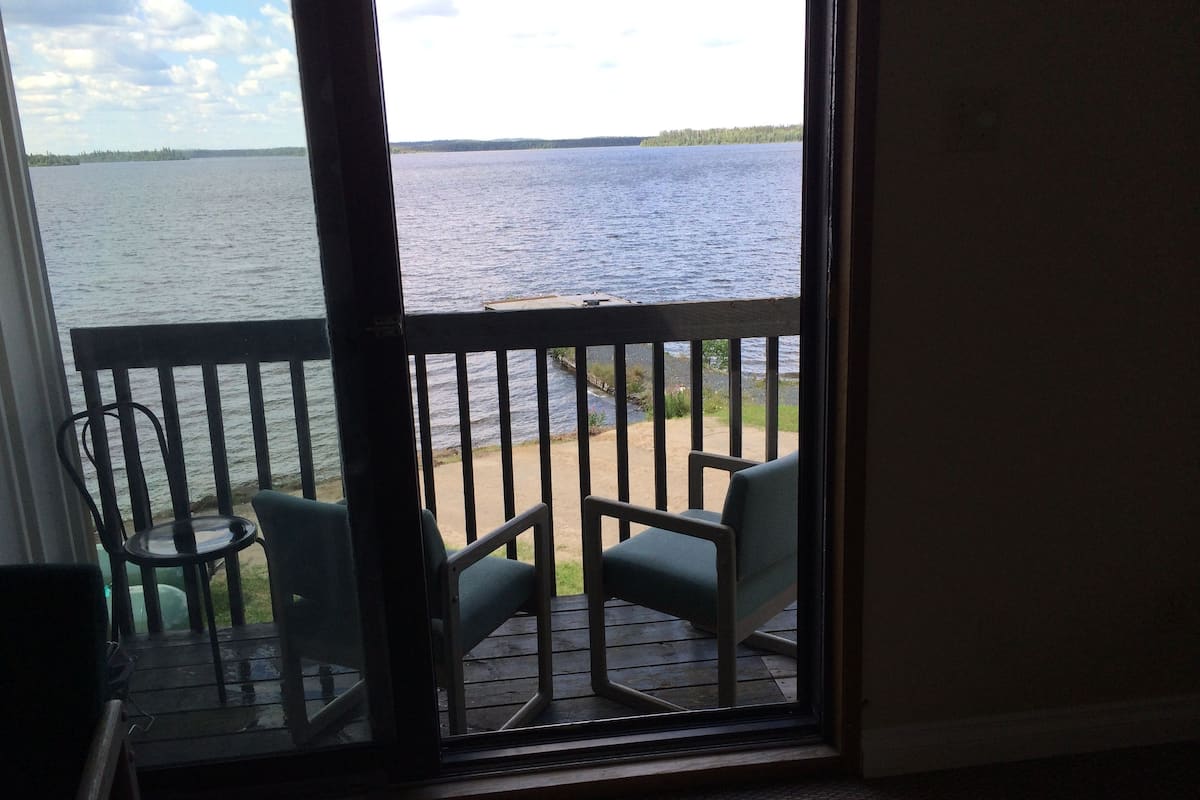 A room with a view of a lake through a window. There are two chairs on a wooden deck outside.