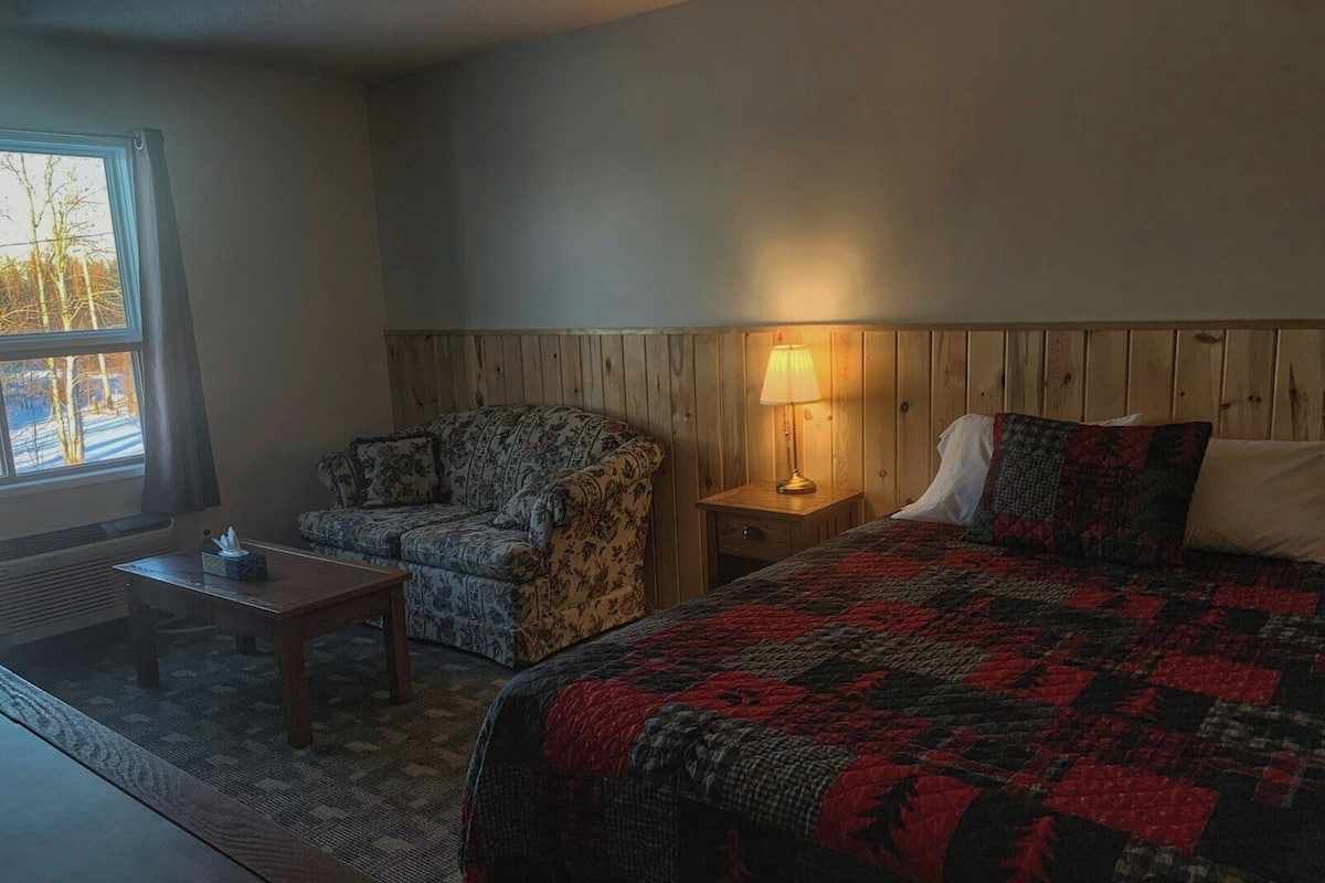 A room with a bed, a chair, a nightstand with a lamp, a small table, and a window with a view of trees.