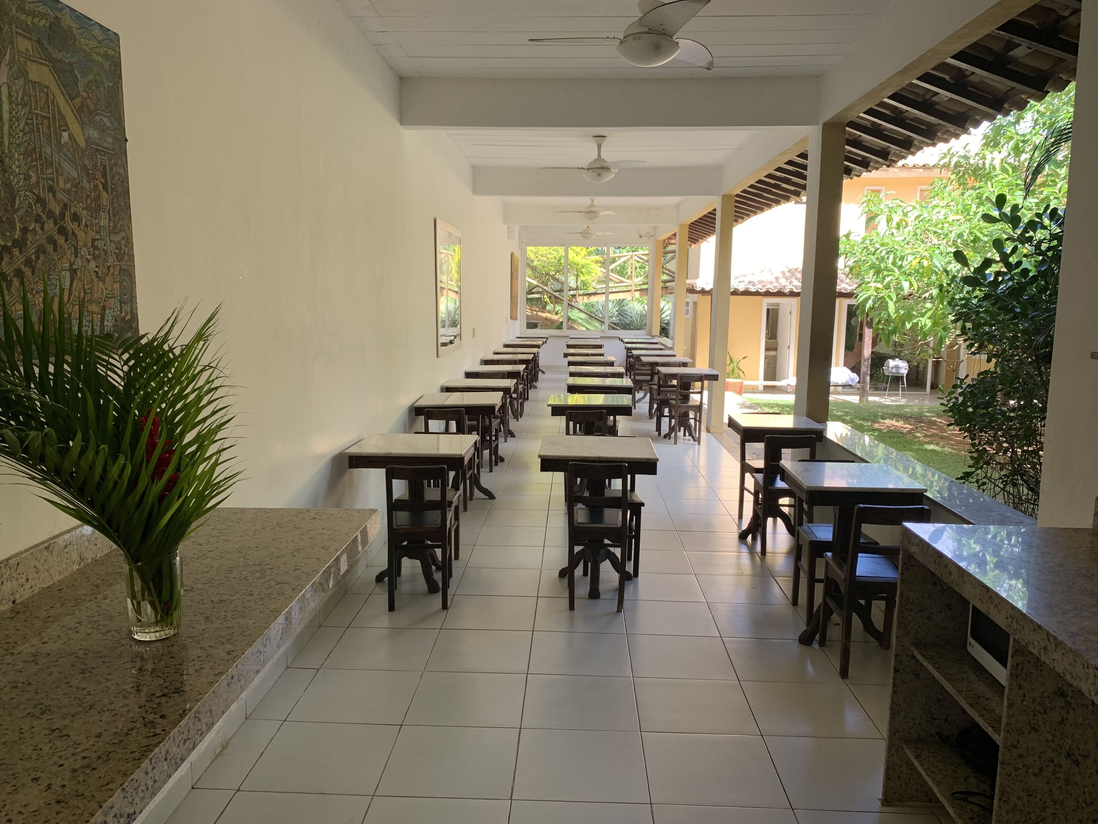 Un long couloir bordé de rangées de tables et de chaises, un ventilateur de plafond et une vue sur un jardin avec des arbres et un bâtiment en arrière-plan.