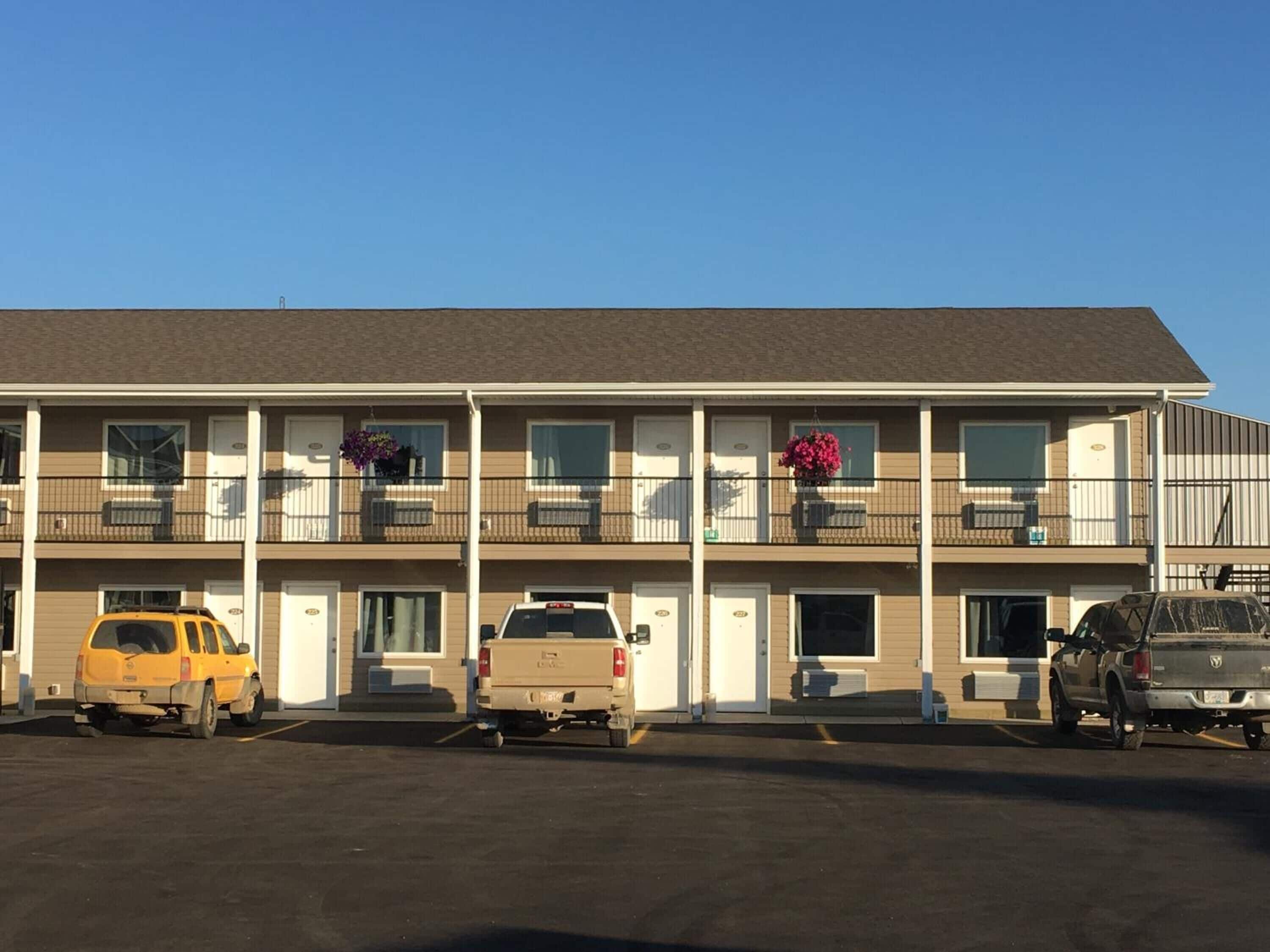 A multi-story motel with a flat roof, multiple balconies with flower baskets, and a parking lot with several vehicles.