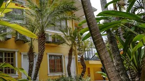 A yellow building with white-framed windows, surrounded by palm trees and tropical foliage. - Palms Hotel Trinidad (Arouca)