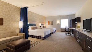 Ein Hotelzimmer mit zwei Betten, einem Sofa, einem Schreibtisch, einem Fernseher und einem Fenster mit Vorhängen.