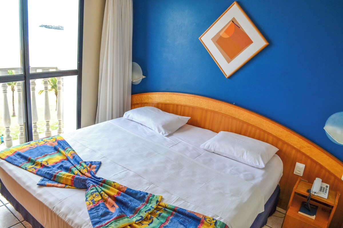A hotel room with a large bed, white linens, a colorful blanket, a wooden headboard, a framed picture on the wall, and a nightstand with a phone.