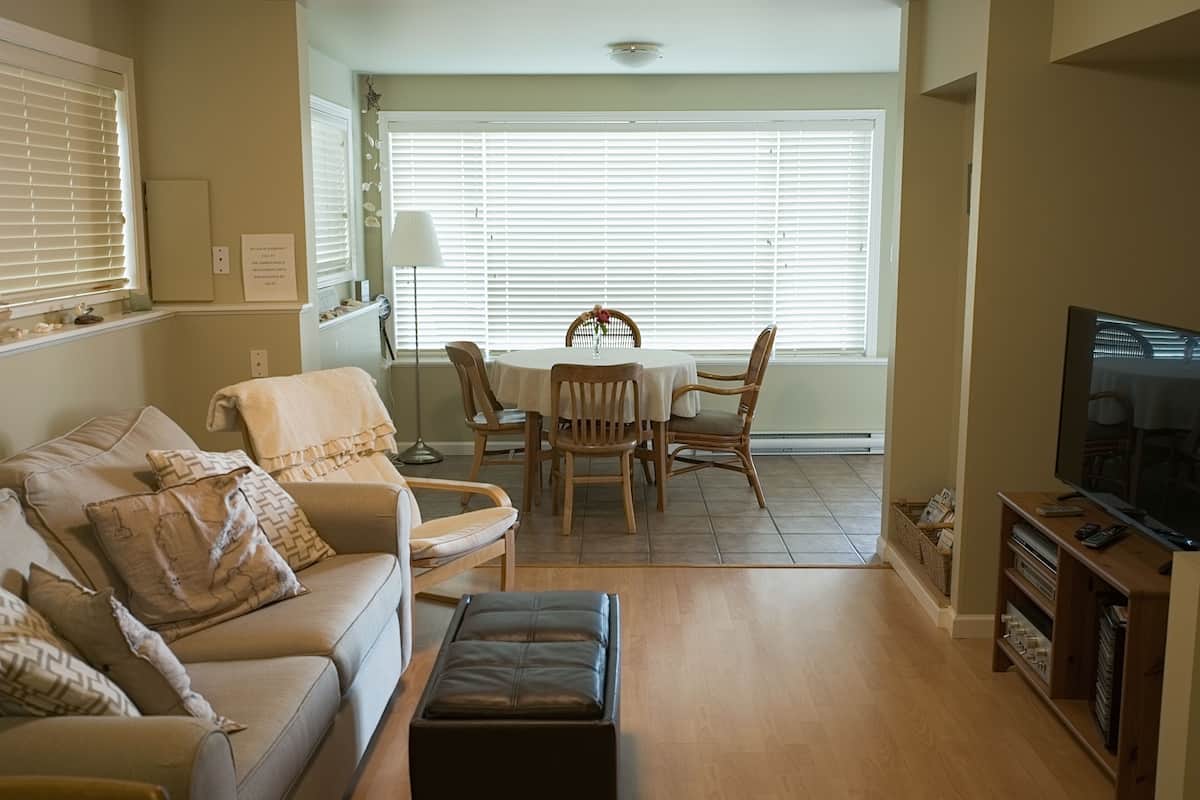 A living room with a sofa, ottoman, and a dining table with chairs. There is a TV stand with a TV and a lamp.