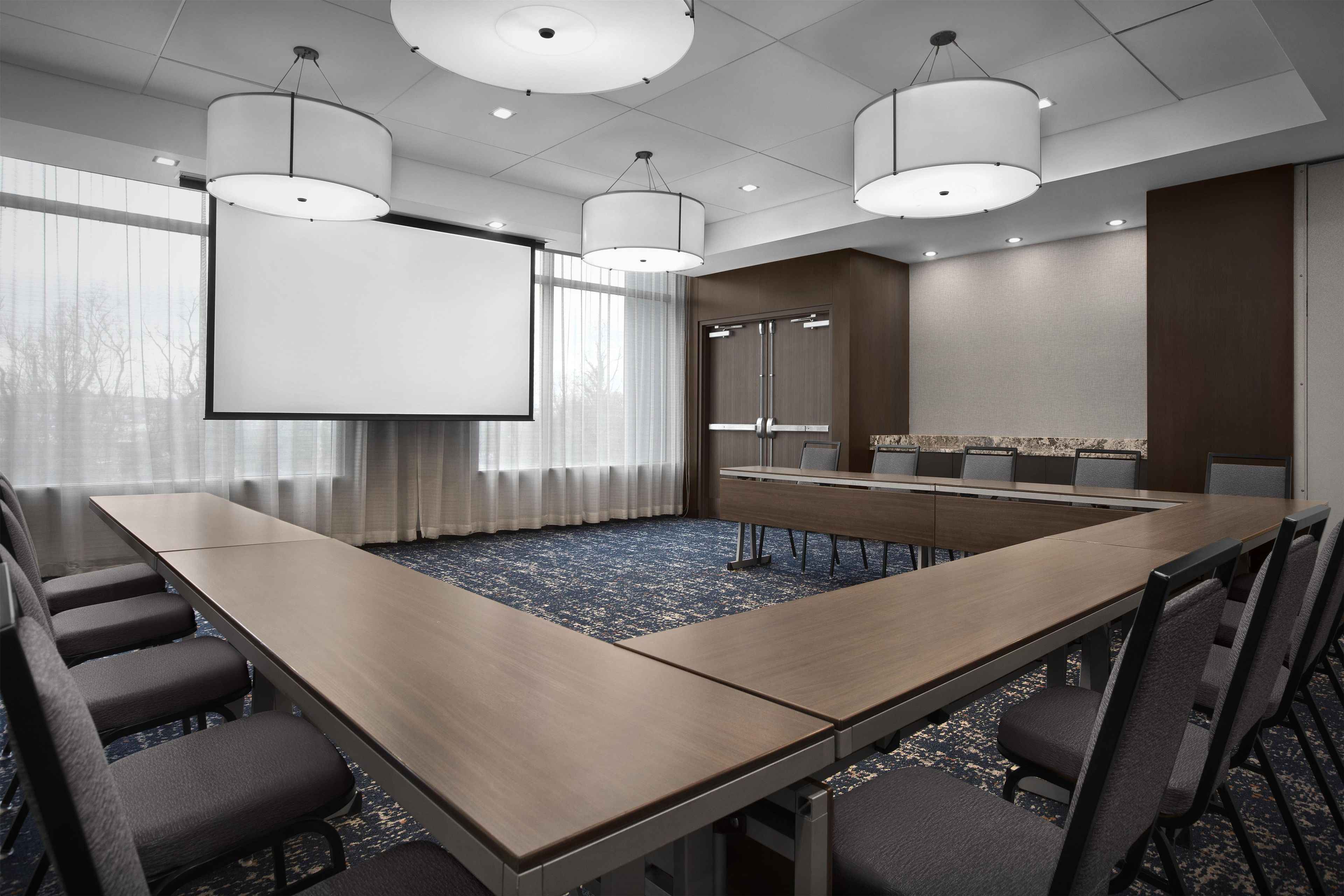 Une salle de conférence équipée d’une longue table, de chaises, d’un écran de projection et d’une grande fenêtre avec des rideaux.