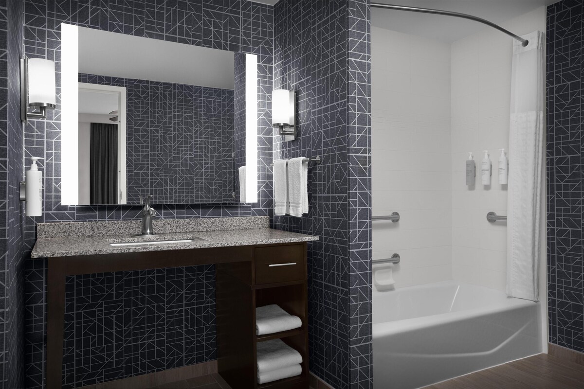 Ein modernes Badezimmer mit dunklem, gemusterten Fliesenrückwand, einer Granit-Arbeitsplatte, einem integrierten Waschbecken, einem großen Spiegel und einer großzügigen Dusche.
