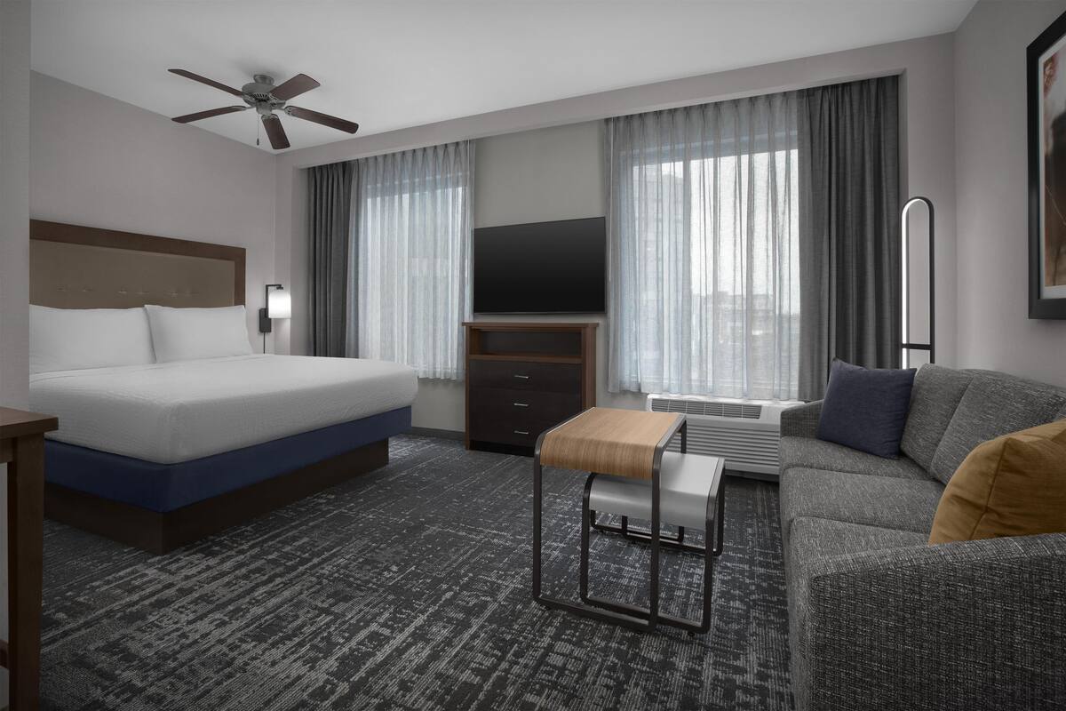 Habitación de hotel con cama, sofá, televisión, ventilador de techo y ventana con cortinas.