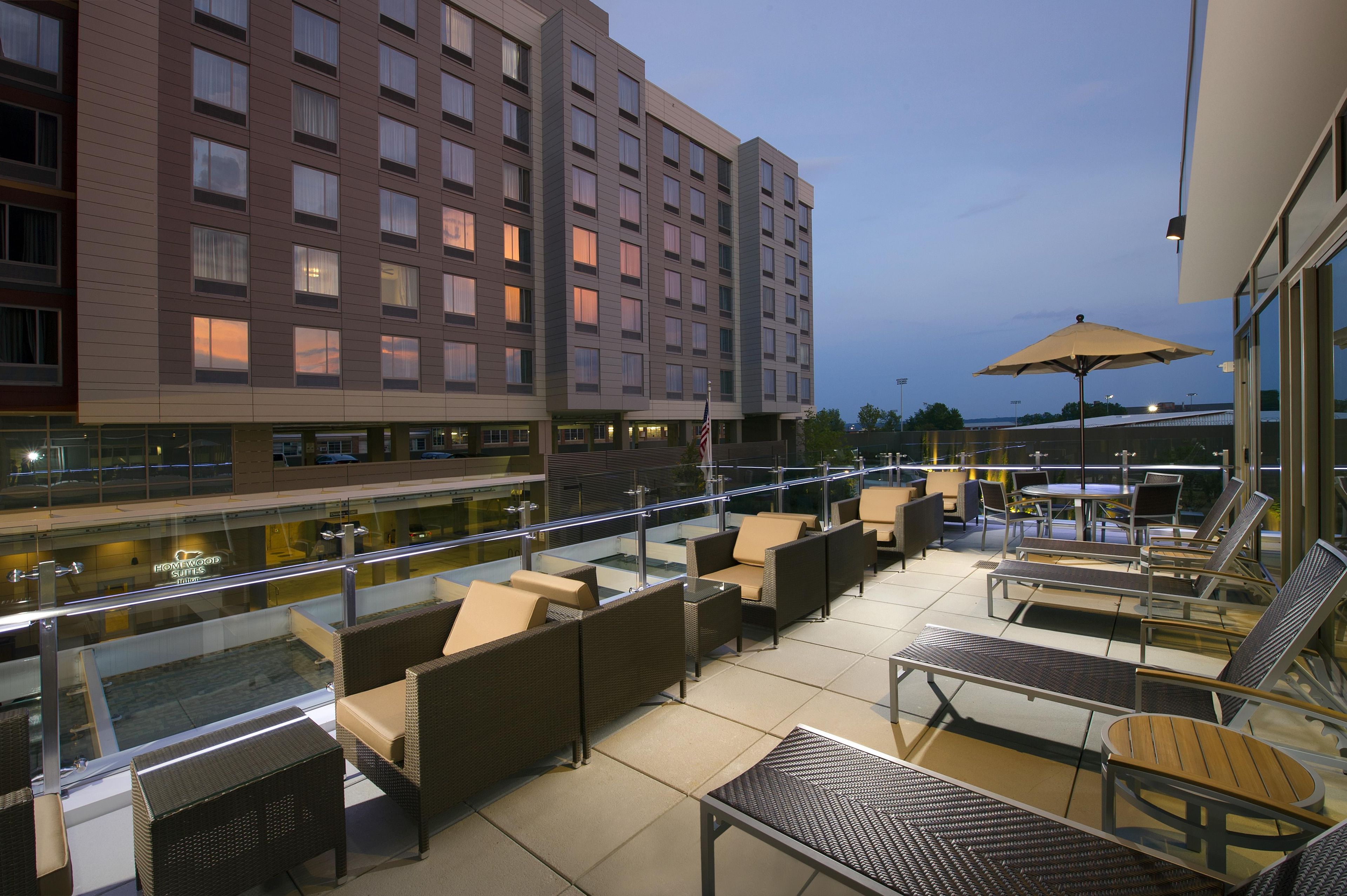 Eine Hotel-Dachterrasse mit Außenmöbeln, einer Glasbrüstung und Blick auf das Hotelgebäude.