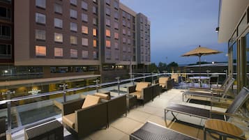 Eine Hotel-Dachterrasse mit Außenmöbeln, einer Glasbrüstung und Blick auf das Hotelgebäude.