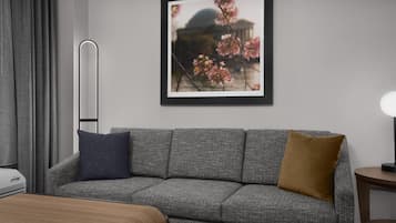 Ein modernes Hotelzimmer mit einem grauen Sofa, einem gerahmten Bild mit Kirschblüten, einem Nachttisch mit Lampe und einem Fenster mit grauen Vorhängen.