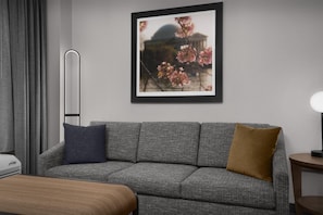 Une chambre d’hôtel moderne avec un canapé gris, un tableau encadré représentant des fleurs de cerisier, une table de chevet avec une lampe et une fenêtre avec des rideaux gris.