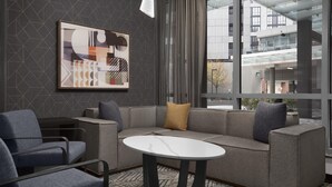 Eine moderne Hotelhalle mit einer grauen Couch, blauen Sesseln und einem weißen Tisch. Ein großes abstraktes Gemälde ziert die Wand, und der Raum verfügt über einen gemusterten Teppich und große Fenster mit Blick auf das Hotelgebäude.