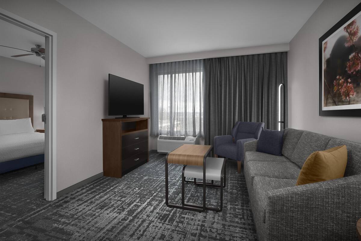 Una habitación de hotel moderna con una cama, un televisor, un sofá, una mesita y un ventanal grande con cortinas.