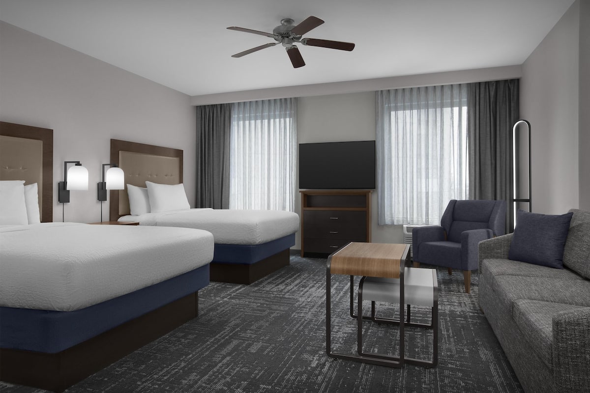 Habitación de hotel con dos camas, televisión, ventilador de techo, escritorio y sofá.