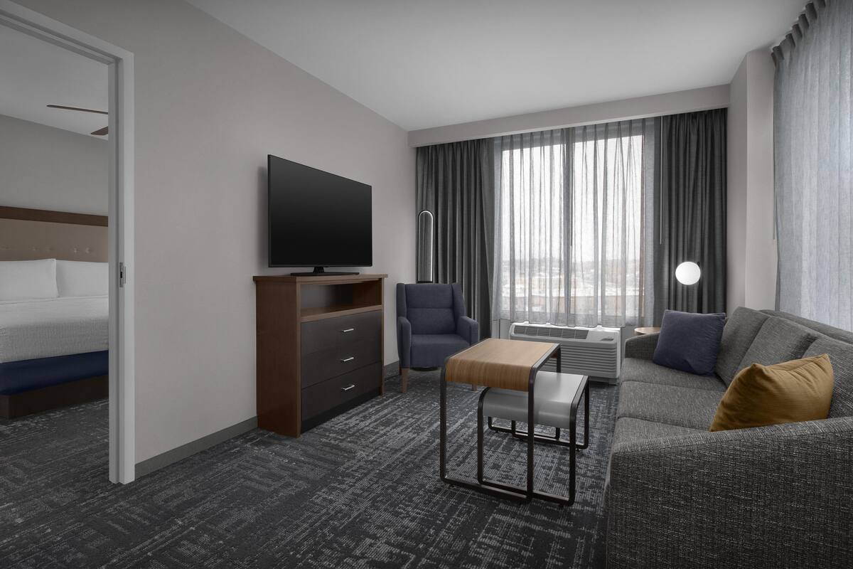 Una habitación de hotel moderna con cama, sofá, televisor, escritorio y un ventanal con cortinas.