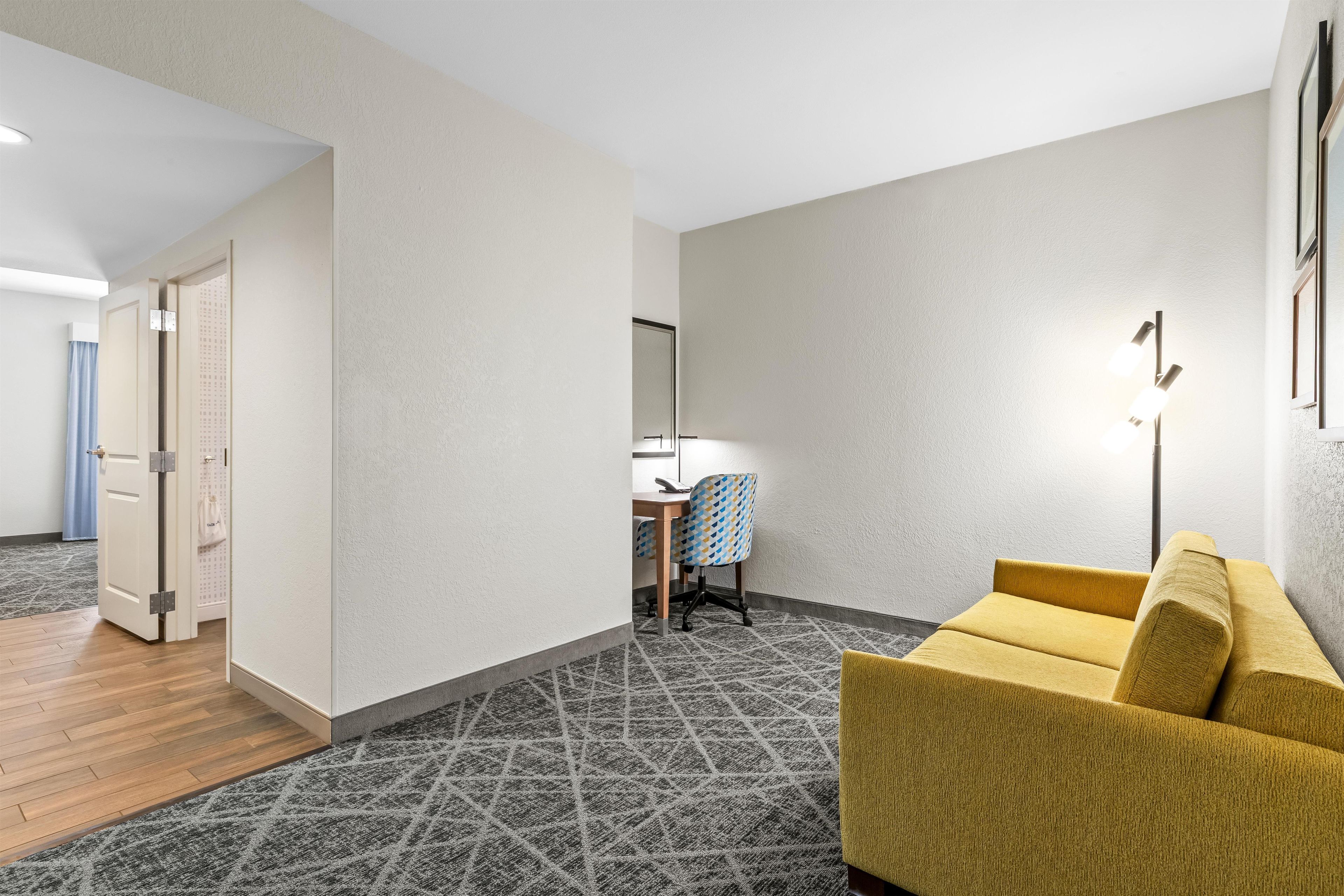 Habitación de hotel con piso de madera, alfombra estampada, sofá amarillo, escritorio con silla y ventana con persianas.