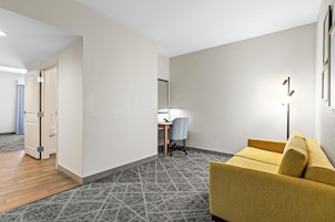 Habitación de hotel con piso de madera, alfombra estampada, sofá amarillo, escritorio con silla y ventana con persianas.
