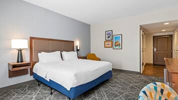 A hotel room with a large bed, a bedside lamp, a small table, and a chair. There are framed pictures on the wall and a door leading to another room.