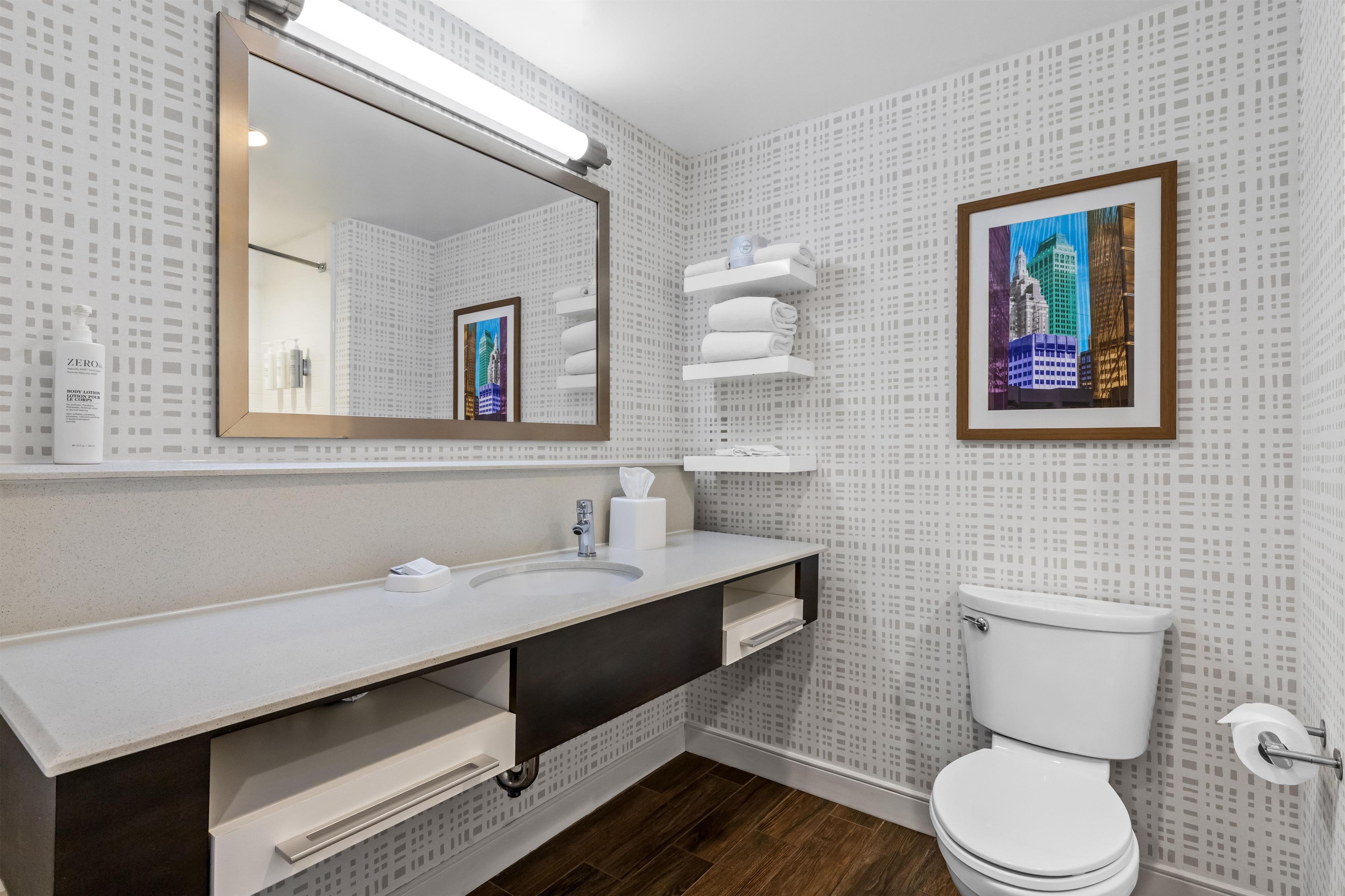 Un baño moderno con un espejo grande, un lavamanos blanco, un inodoro y una fotografía enmarcadas de un paisaje urbano en la pared.