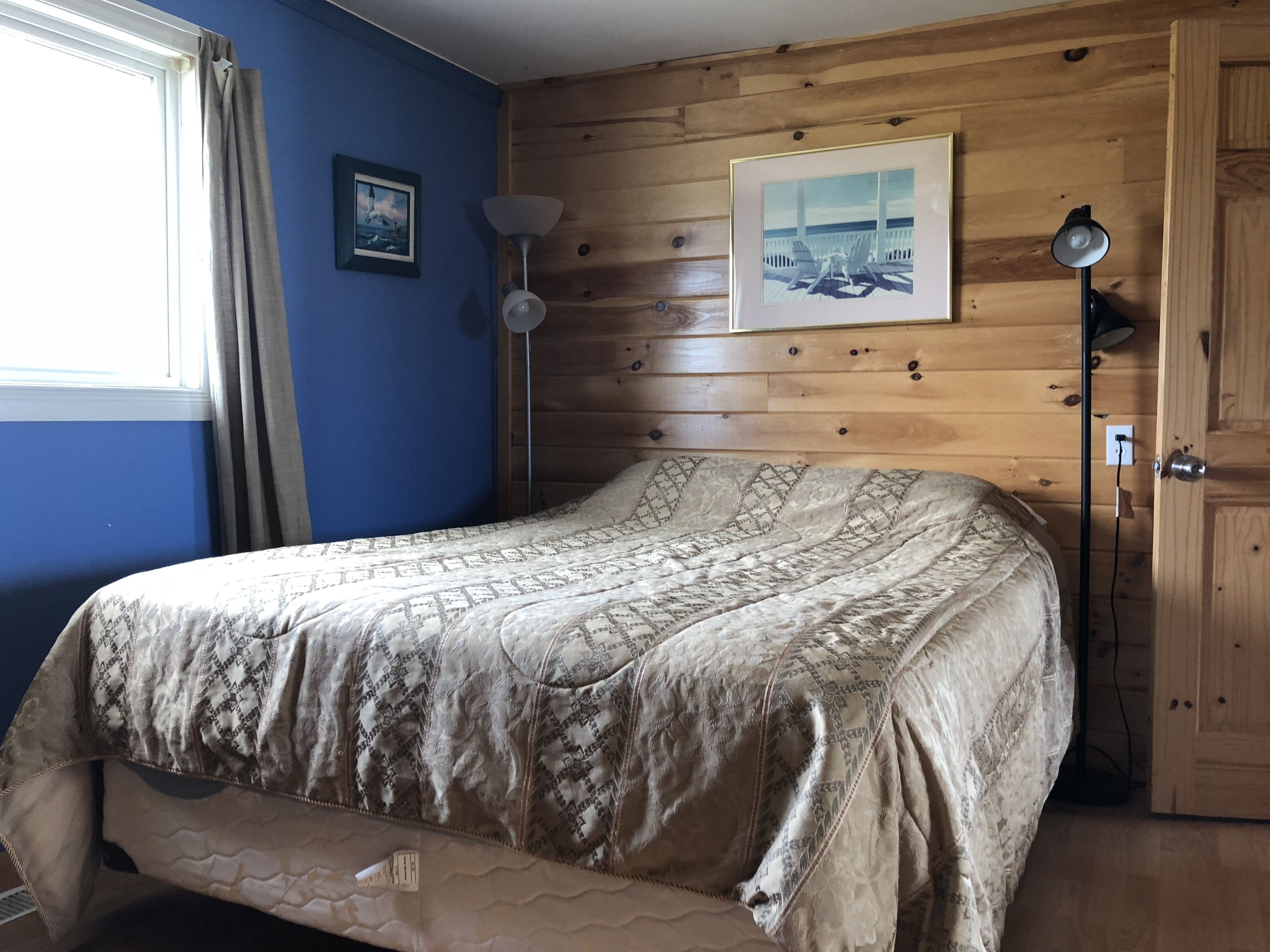 Un dormitorio con cama, cabecero de madera, un cuadro enmarcado, una lámpara de pie y una puerta.