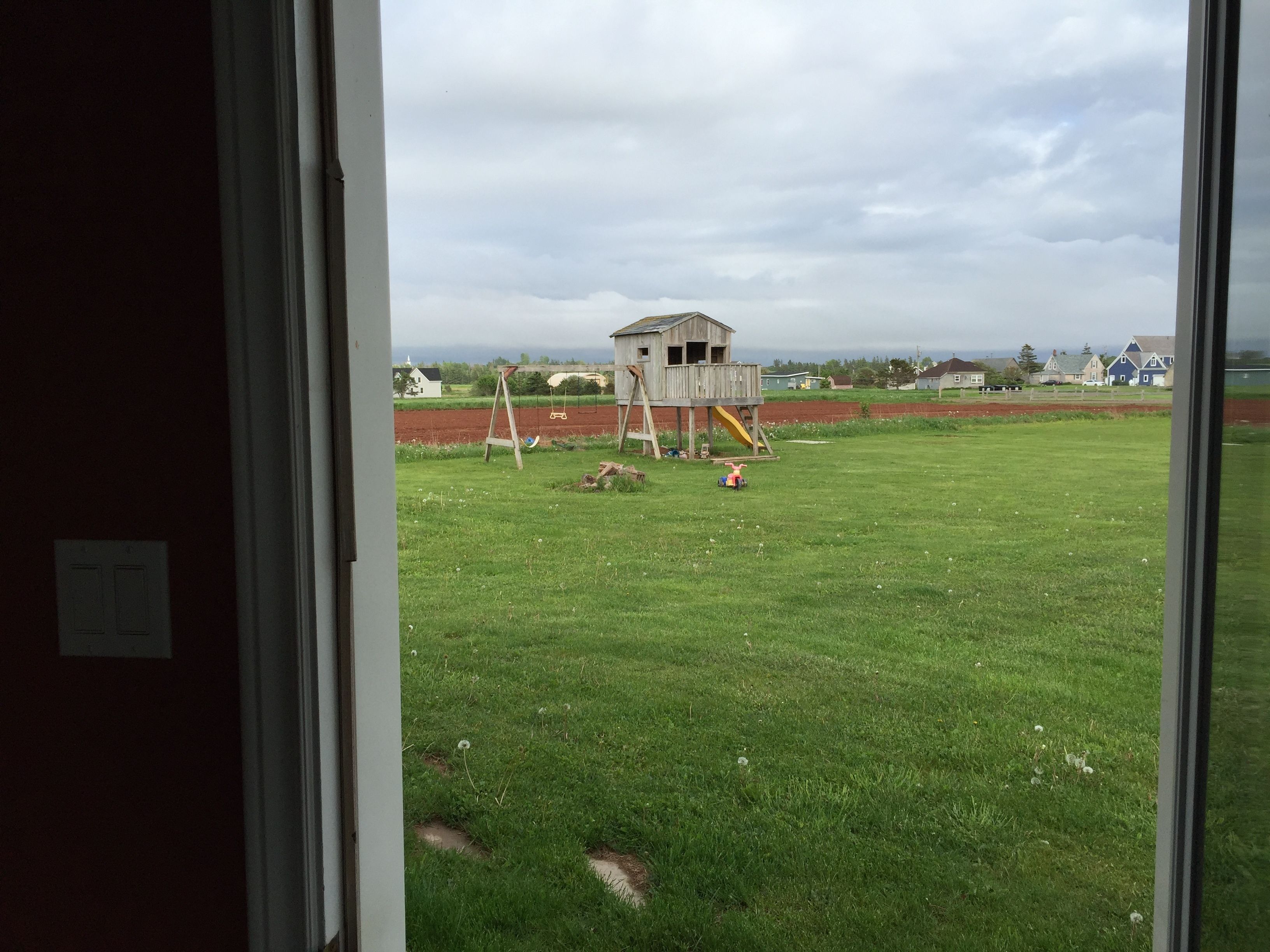 A view through a window of a grassy field with a wooden playhouse and swings.