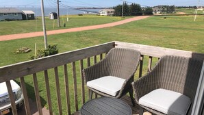 A wooden deck with two wicker chairs and a small table, overlooking a grassy yard with a path and distant houses.