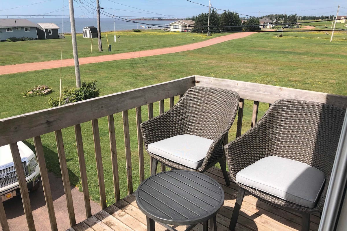 Una terraza de madera con dos sillas de mimbre y una mesita, con vistas a un jardín con un sendero y casas a lo lejos.