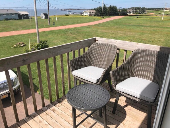 A wooden deck with two wicker chairs and a small table, overlooking a grassy yard with a path and distant houses.